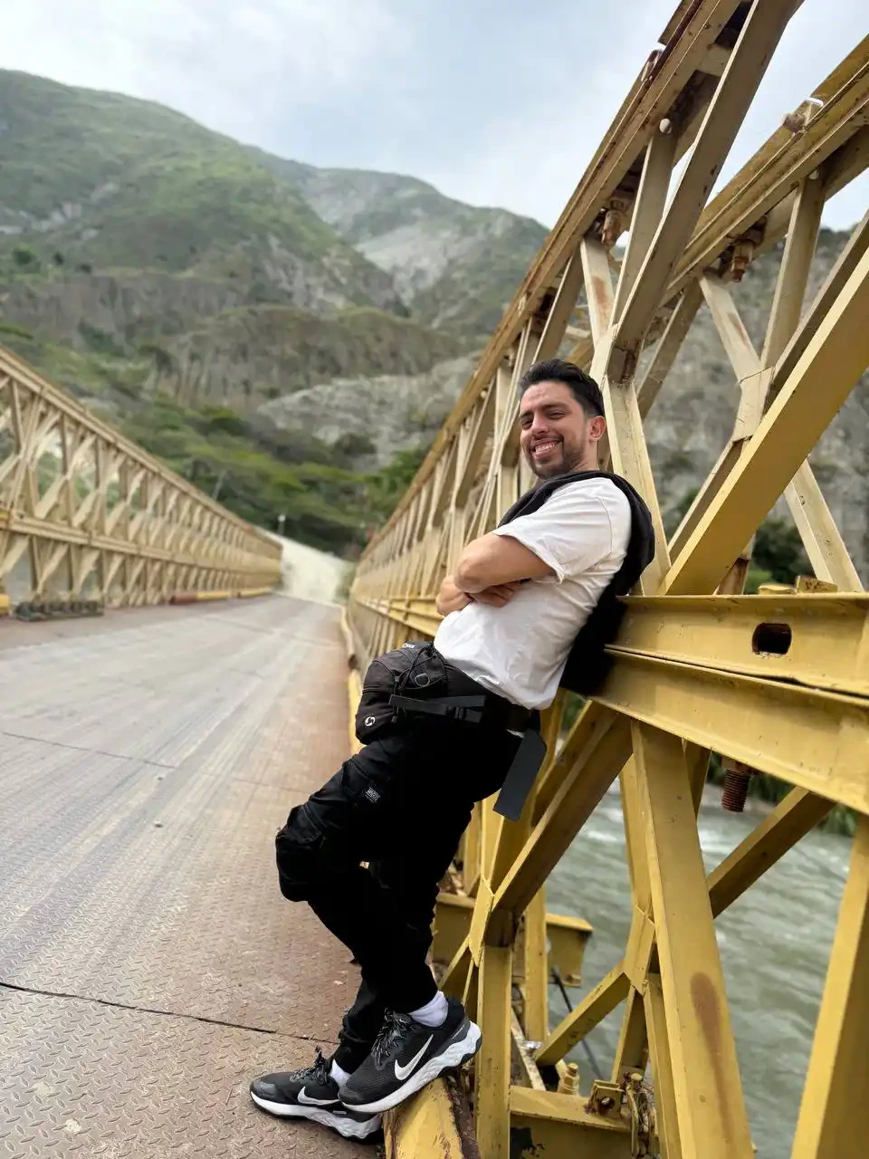 Rafael Cuevas smiling, leaning on the steel truss of a yellow bridge over a mountain river